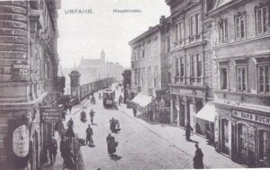 Blick aus der Hauptstraße in Urfahr um die Zeitenwende vom 19. ins 20. Jhdt. auf den Brückenkopf und Linz. Eine "Elektrische" (Straßenbahn) bahnt sich ihren Weg durch geschäftiges Leben und Treiben. Im Hintergrund die Ignatiuskiche in Linz, heute als "Alter Dom" bekannt. Foto: Sammlung oepb